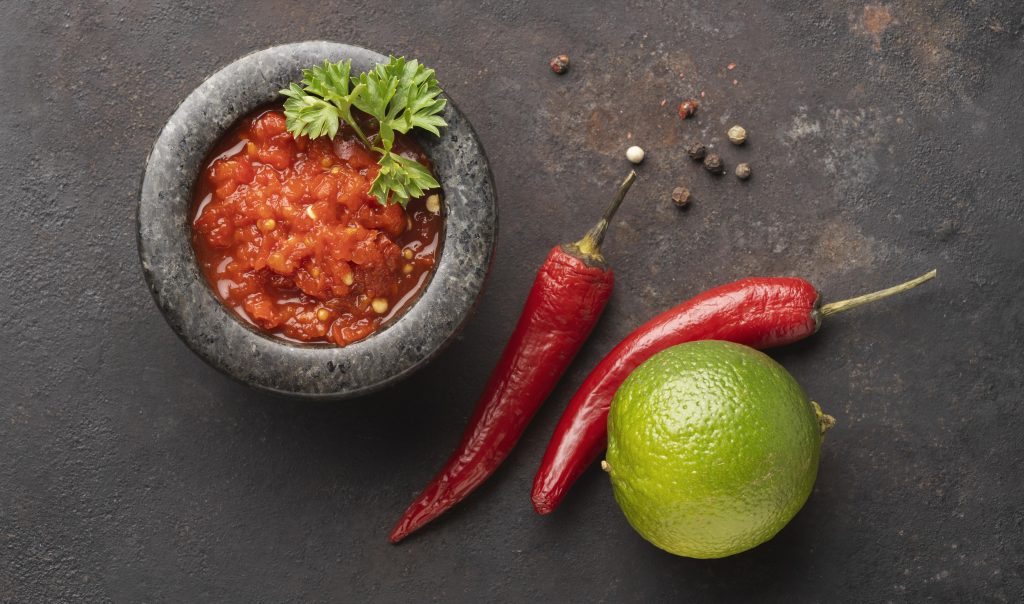 Composition with Mexican molcajete filled with salsa, chili pepper, and lime illustrates blog "Are Carnitas Spicy?"